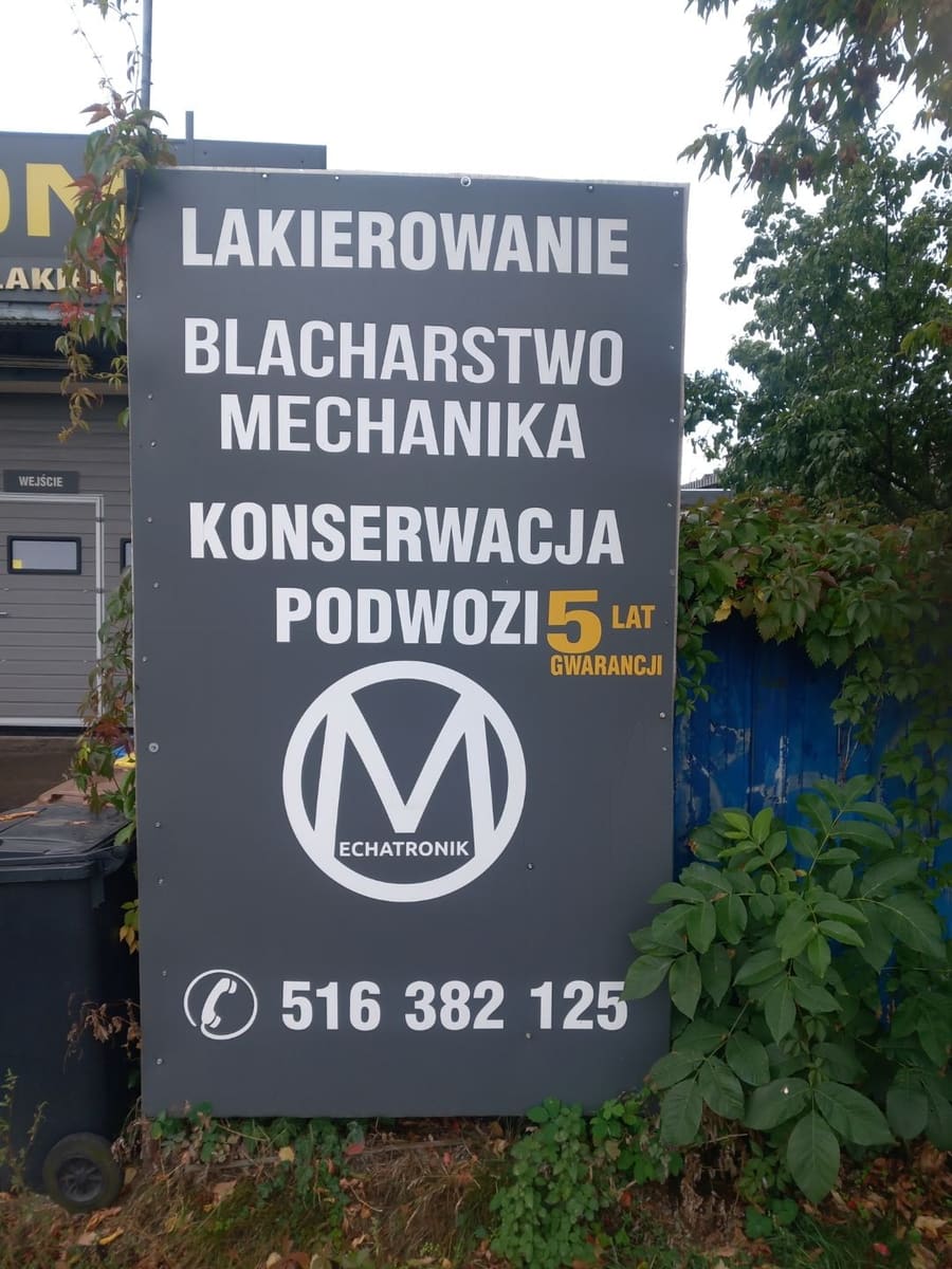 Tablica reklamowa warsztatu Mechatronik z napisem lakierowanie, blacharstwo, mechanika, konserwacja podwozi — 5 lat gwarancji, logo warsztatu i numer telefonu 516 382 125.